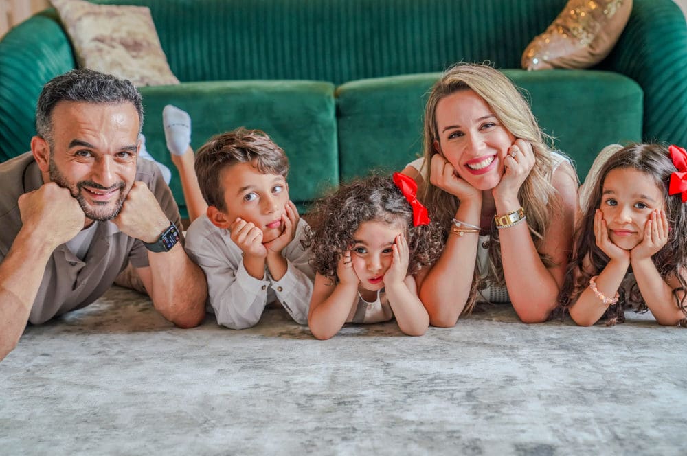 A family lies on the carpet in front of a green couch, posing with their heads resting on their hands. Among them are Dr. Fateh Arslan and Dr. Ayla Idilbi, orthodontics experts from Fiesta Orthodontics, joined by three joyful children, creating a heartwarming scene of togetherness in San Antonio, TX.