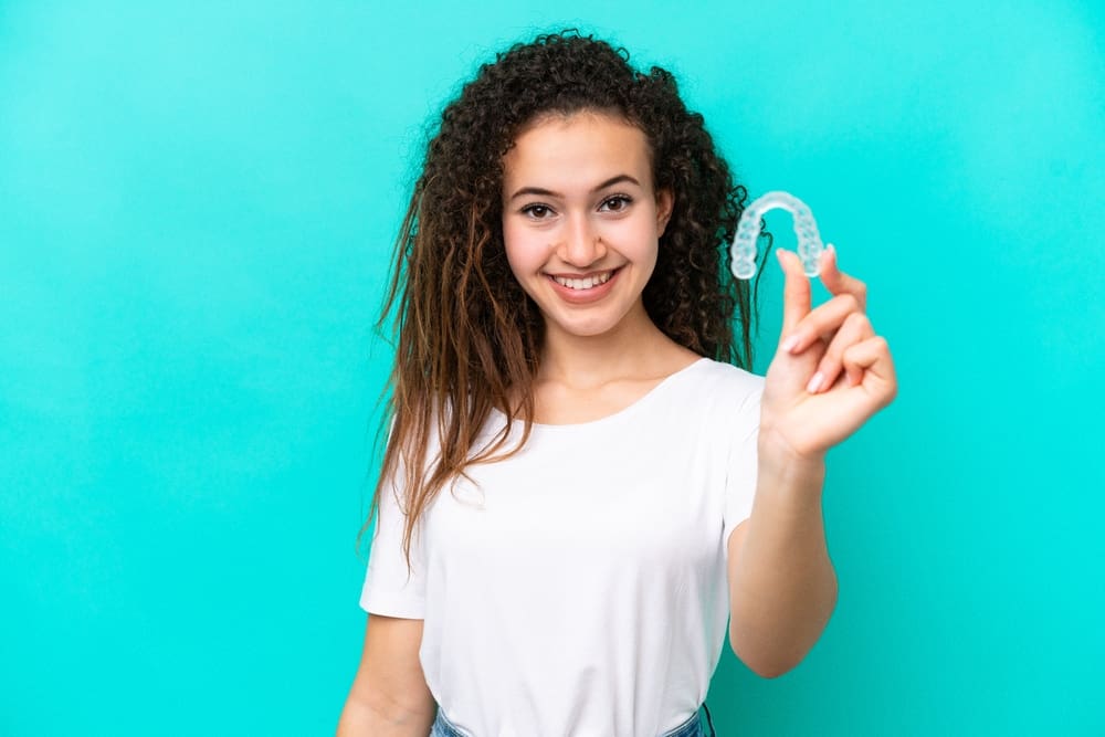 Person in a white shirt holding a clear dental aligner, an excellent alternative to braces, against a turquoise background, smiling at the camera—showcasing innovative orthodontic solutions available in San Antonio, TX.