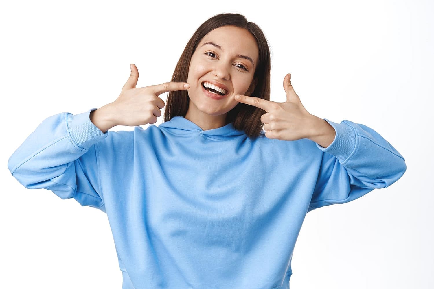A woman in a blue hoodie pointing at her smiling mouth, showcasing her metal braces against a white background in San Antonio, TX