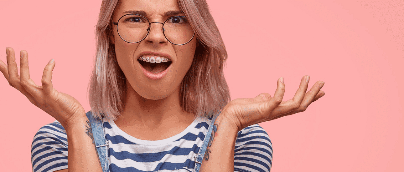A person with glasses and braces, showcasing the benefits of adult orthodontics, wears a striped shirt with an open-mouthed expression and raised hands against a pink background in San Antonio, TX
