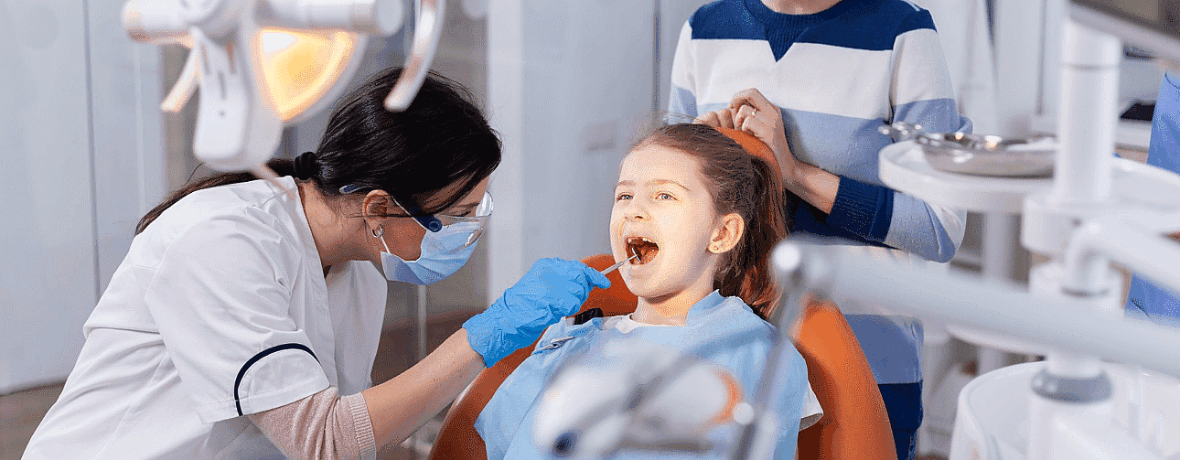 At Fiesta Orthodontics, an orthodontic expert in San Antonio TX, a dentist examines a young patient's teeth while she sits in a dental chair, with an adult standing nearby in San Antonio, TX