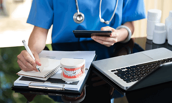 Person in blue scrubs with a stethoscope uses a smartphone and writes on a clipboard, reviewing orthodontic insurance plans. A dental model and laptop are on the desk in San Antonio, TX
