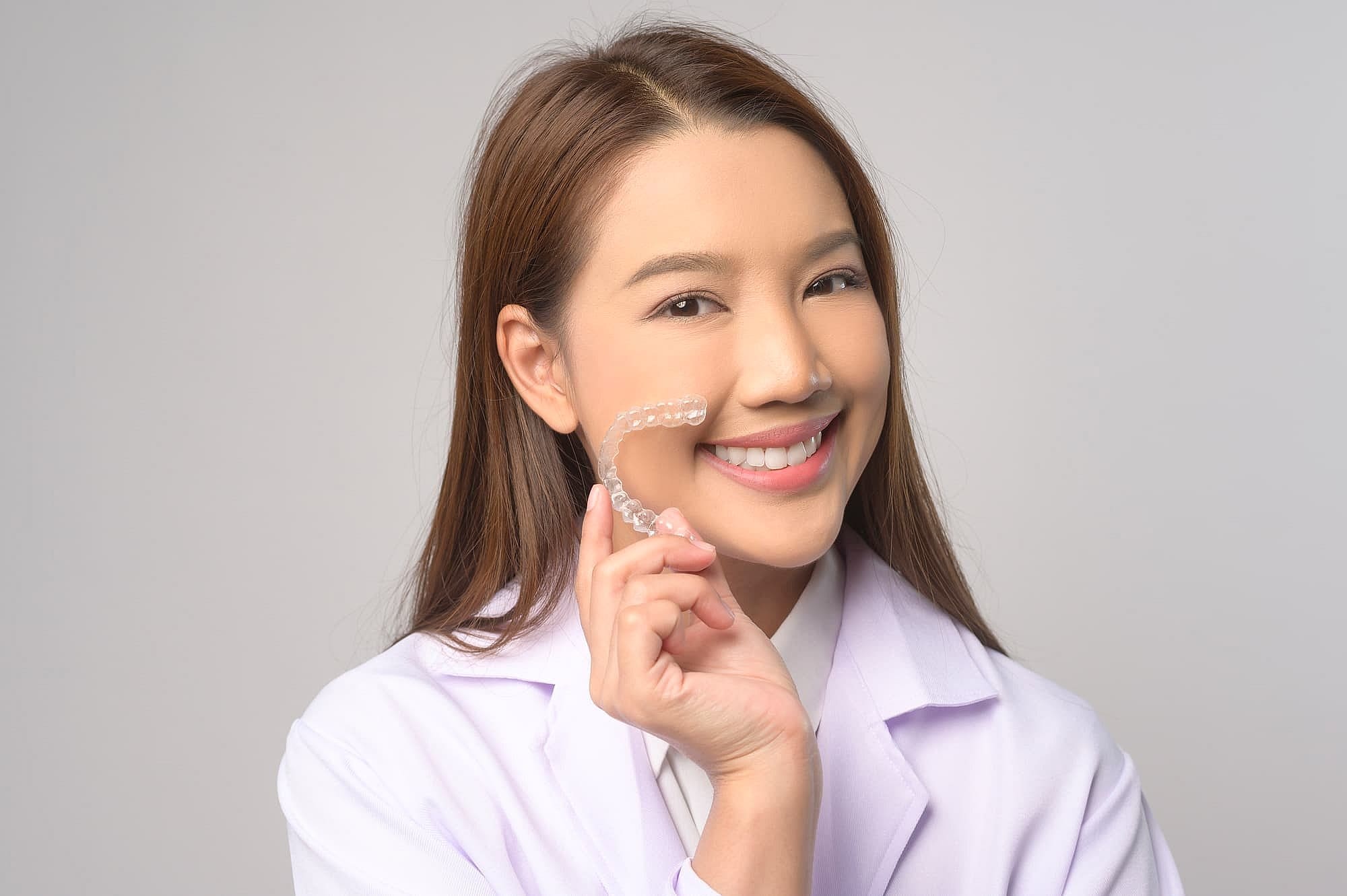 A woman in a white coat holds a clear dental aligner, potentially covered by insurance, and smiles against a plain background in San Antonio, TX