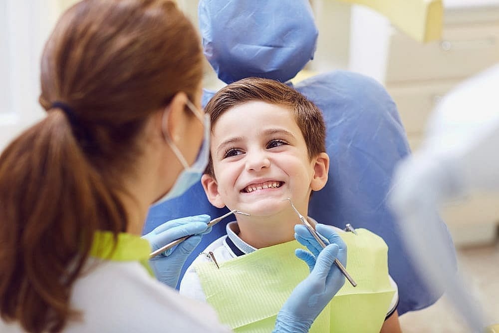 A child with a bright smile sits in the dental chair as the dentist, masked and gloved, gently examines their teeth with care, perhaps discussing the future benefits of a palatal expander in San Antonio, TX