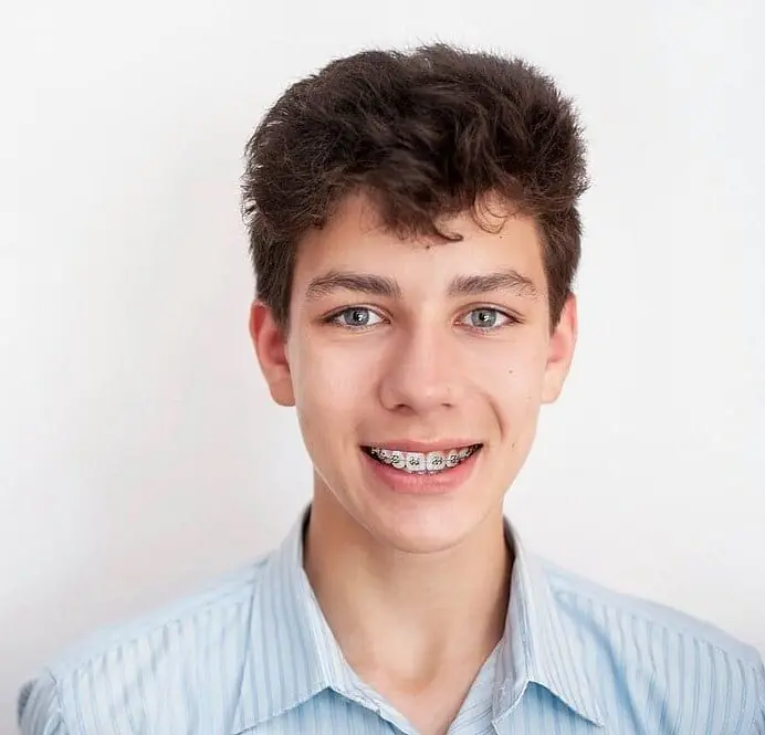 A teenage boy with curly dark hair and braces, showcasing the benefits of early orthodontic care, stands smiling confidently in a light blue striped shirt against a plain white background in San Antonio, TX A teenage boy with curly dark hair and braces, showcasing the benefits of early orthodontic care, stands smiling confidently in a light blue striped shirt against a plain white background in San Antonio, TX