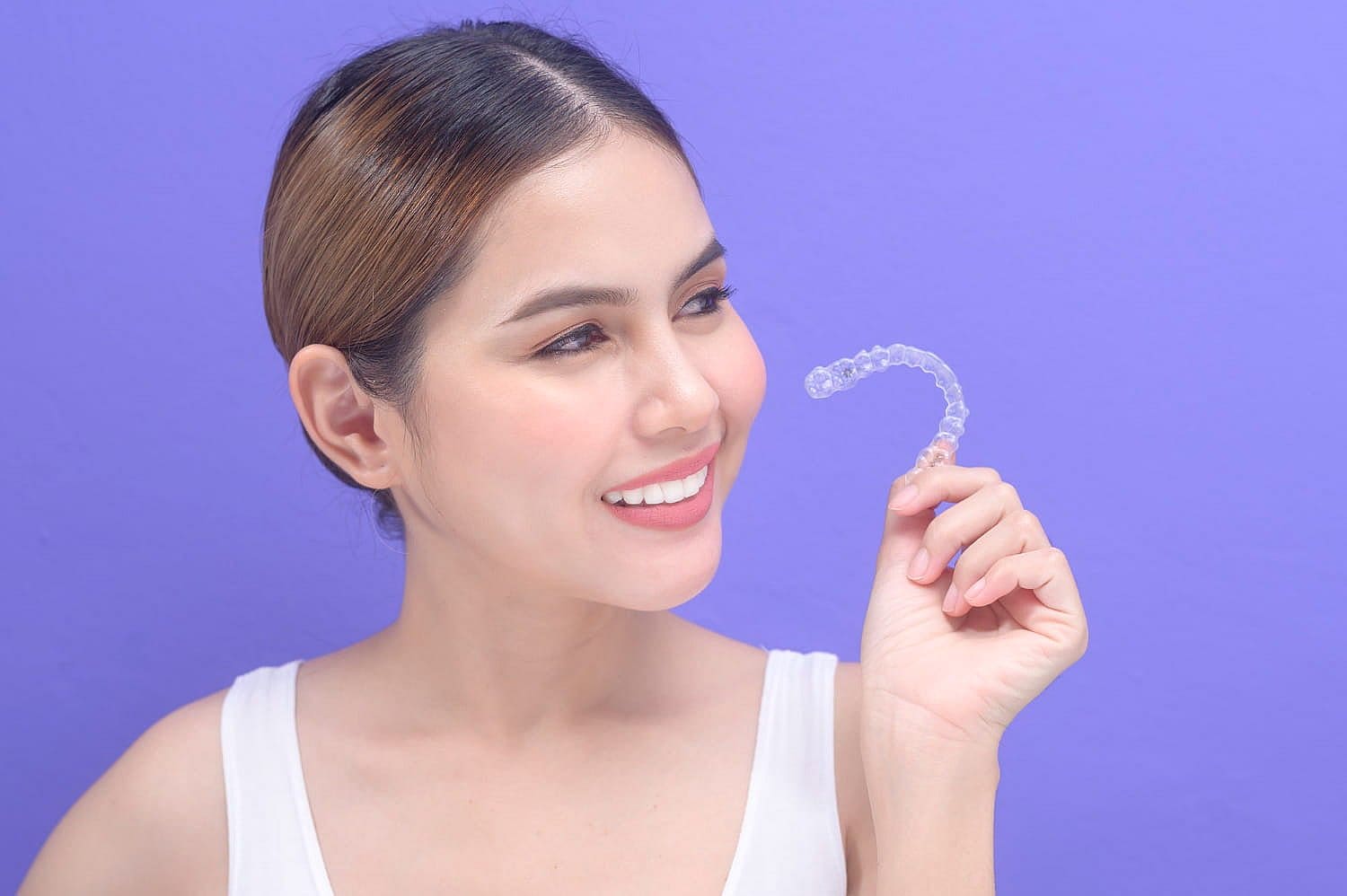 A woman smiles while holding a clear orthodontic aligner against a purple background, showcasing the transformative power of Invisalign. With tailored payment plans, achieving the perfect smile has never been more accessible in San Antonio, TX