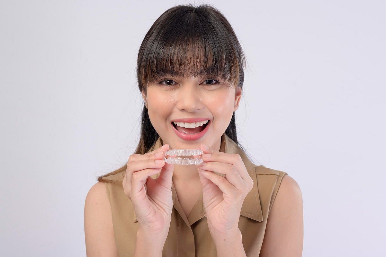 A person, donning a tan sleeveless top, smiles brightly while holding their Invisalign® Clear Aligners as alternative to braces near me against a plain background in San Antonio, TX