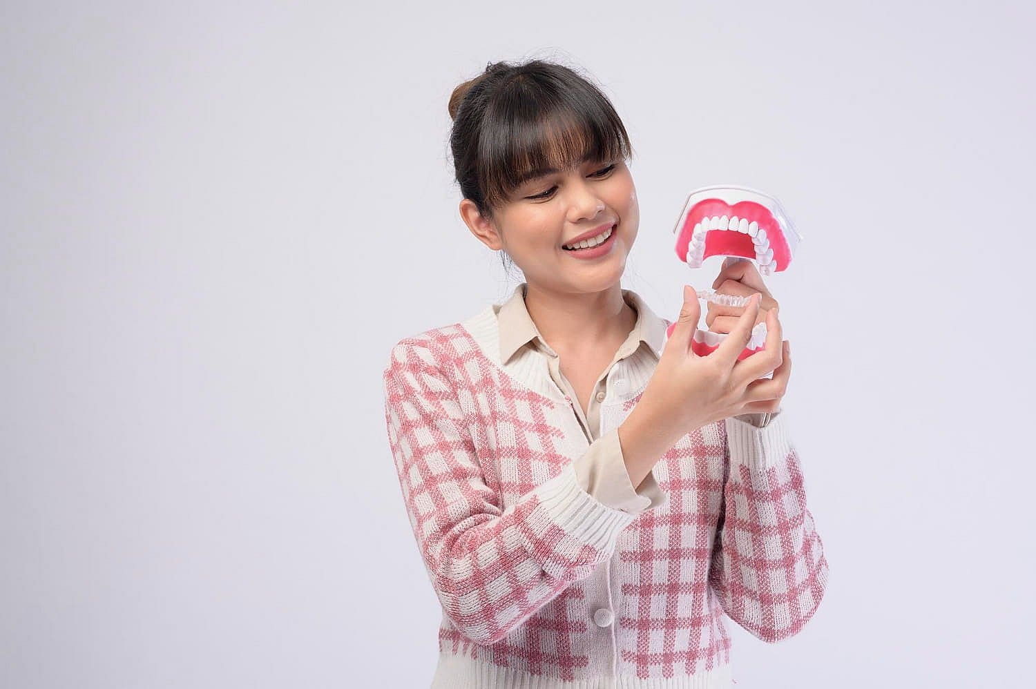 A person wearing a checked cardigan smiles while holding a dental model with visible teeth against a plain background, showcasing the benefits of using Invisalign for that perfect smile in San Antonio, TX