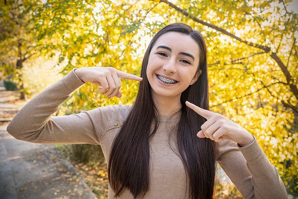 Young woman with long dark hair smiling and pointing to her metal braces, standing outdoors with vibrant yellow leaves in the background in San Antonio, TX