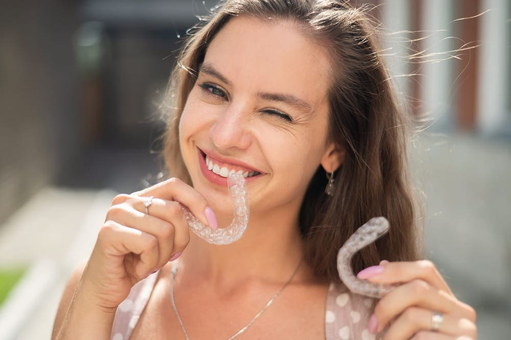 A woman beams and winks while showcasing two clear dental aligners, epitomizing the expertise of a San Antonio TX orthodontist, outdoors.