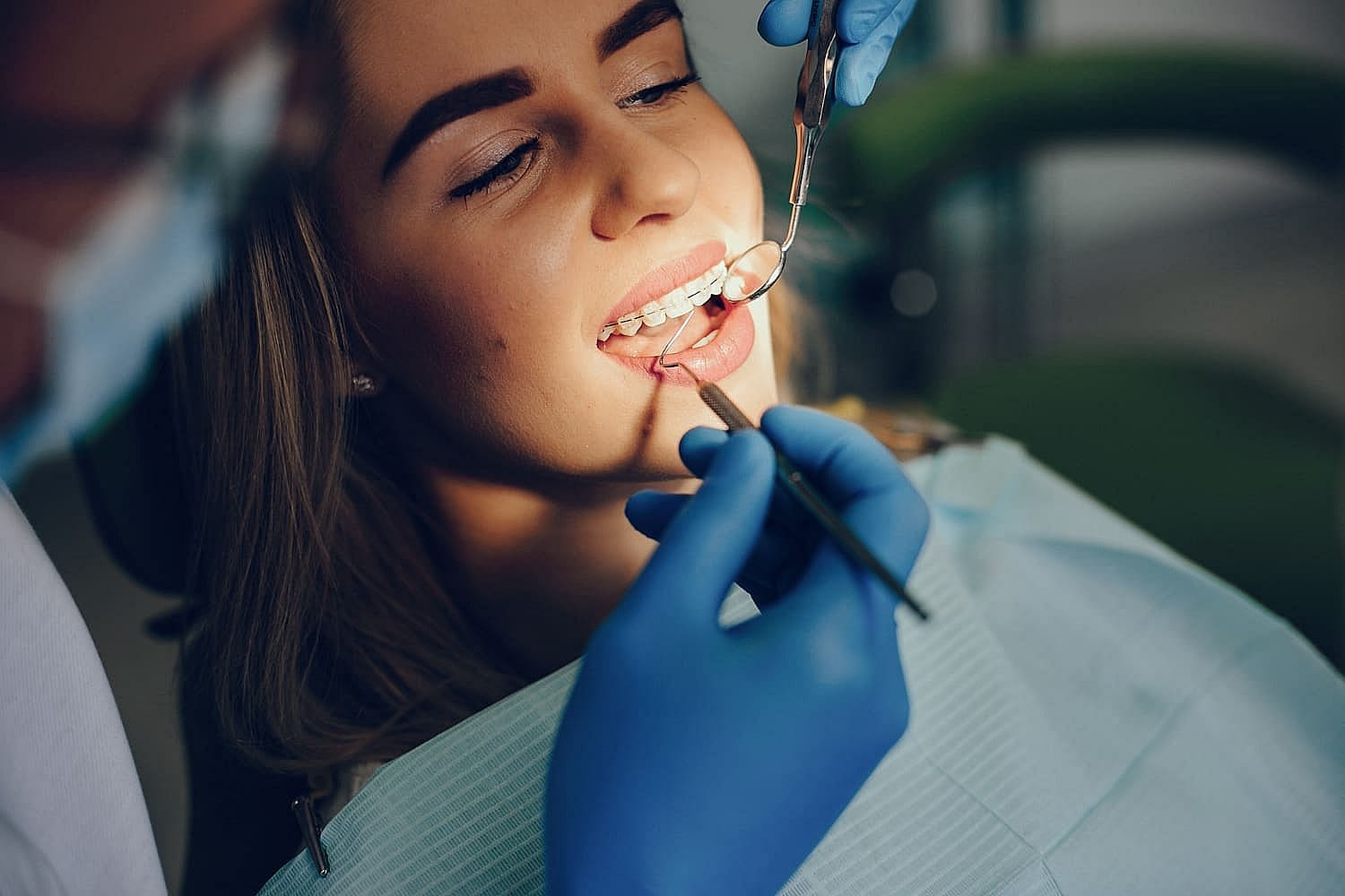 A woman with clear braces near me is sitting in a dental chair, receiving an orthodontic checkup by a dentist wearing blue gloves, holding dental tools in San Antonio, TX