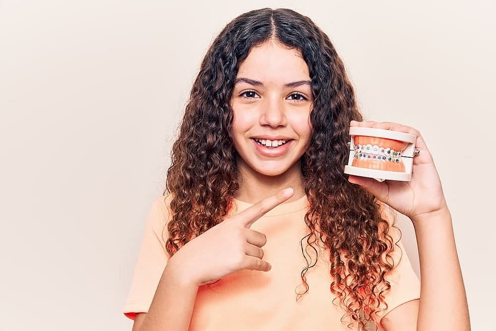 Fiesta Orthodontics A girl with curly hair beams brightly, pointing at a dental model featuring braces, pondering the cost in San Antonio, TX
