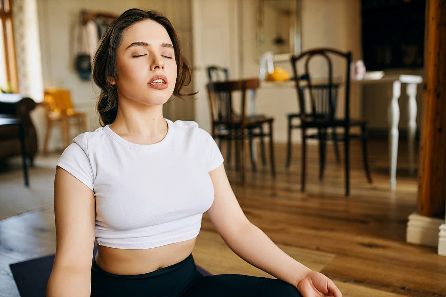 A woman in a white top sits cross-legged on a mat with eyes closed, appearing to meditate in a room with wooden flooring and chairs. While she remains serene, her subtle mouth breather adds an element of calm to the tranquil setting in San Antonio, TX