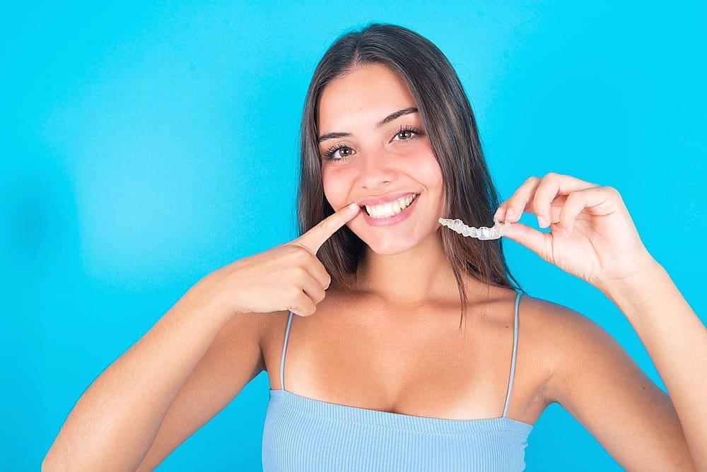 A woman smiles and points to her teeth while holding a clear aligner against a blue background, showcasing the benefits of orthodontic insurance plans in San Antonio, TX