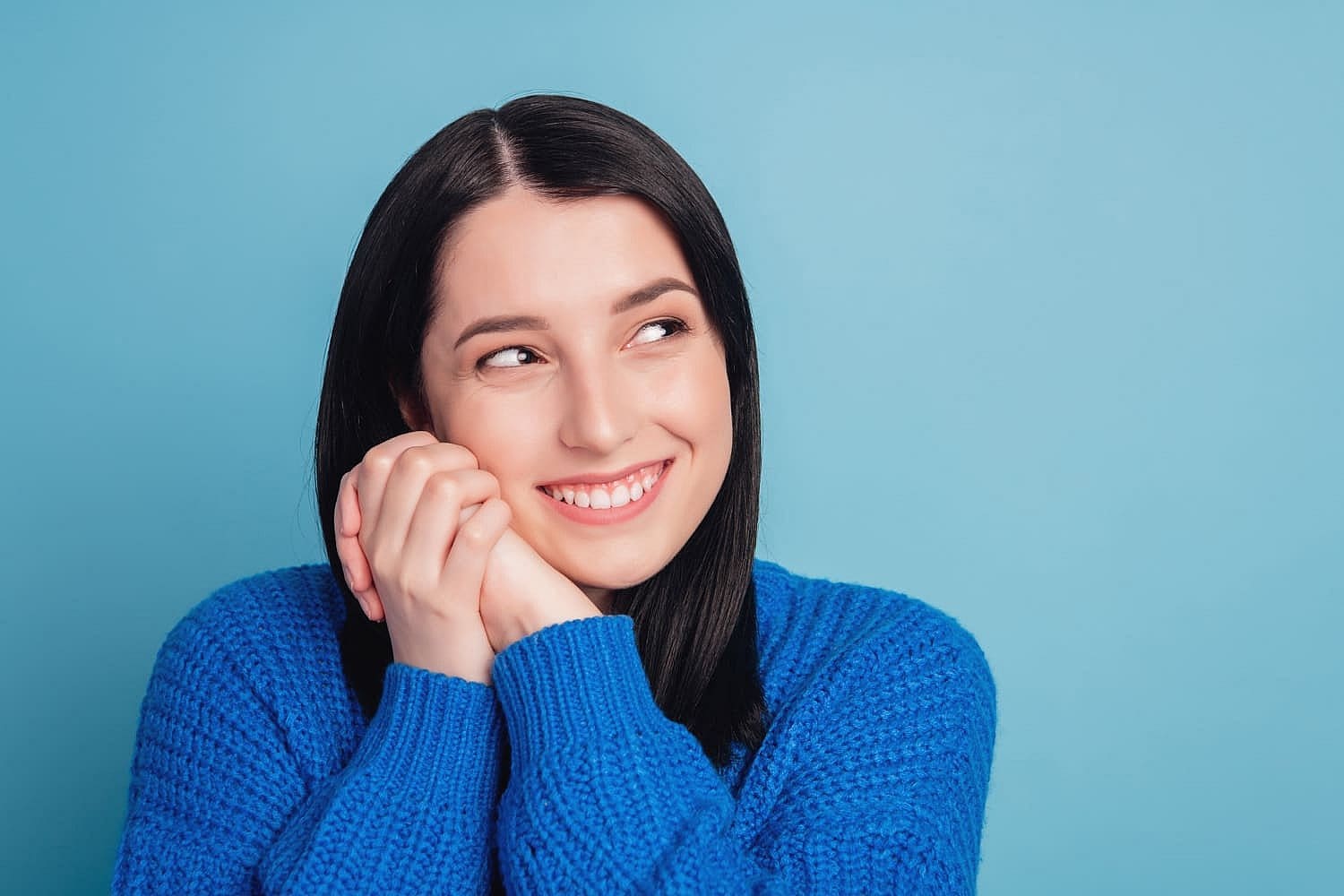A woman with long dark hair, wearing a blue sweater, flashes a bright smile enhanced by Invisalign, as she gazes to the side against a serene blue background in San Antonio, TX