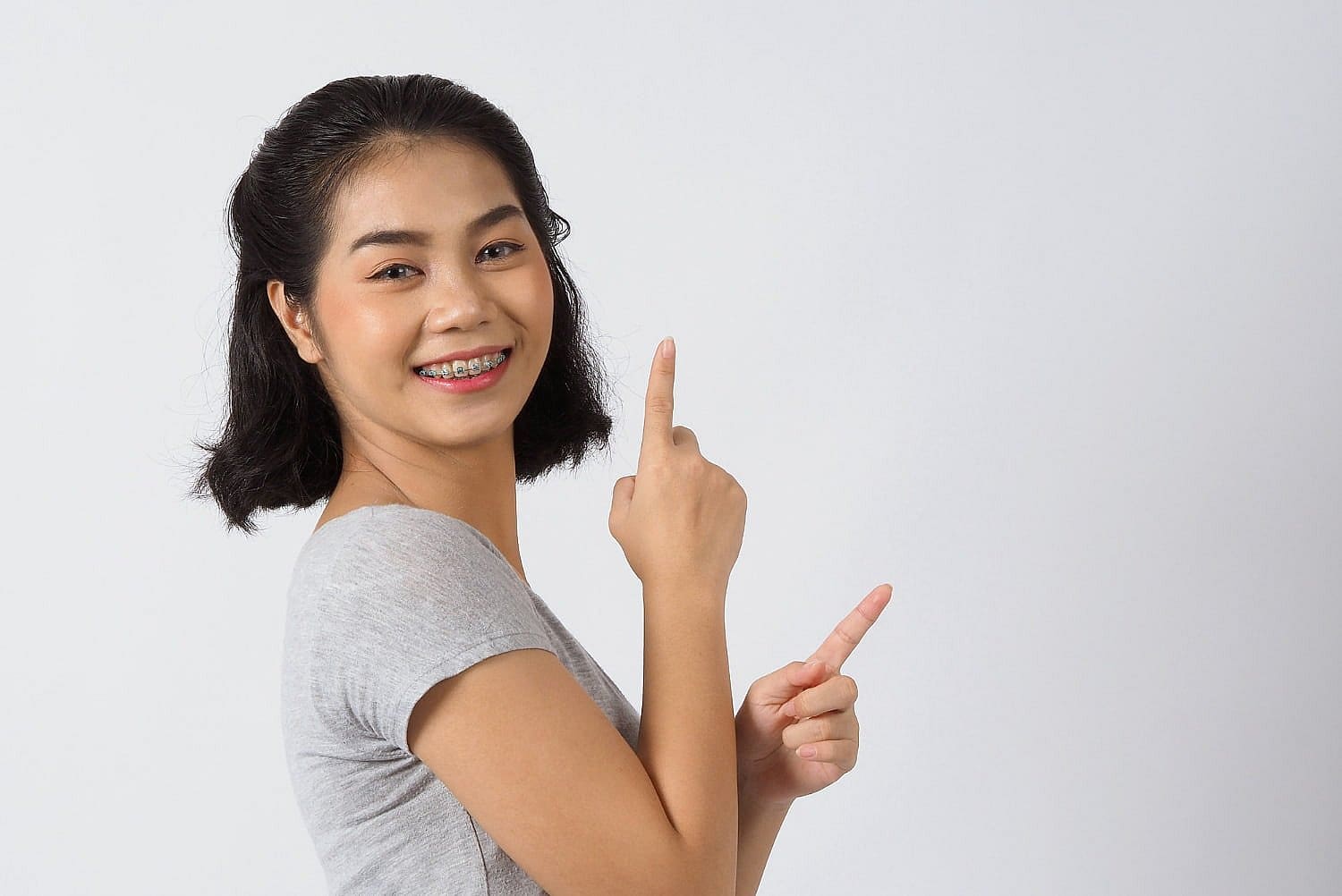 Fiesta Orthodontics A woman with dark hair, wearing a gray shirt, is joyfully pointing upwards with both hands against a plain white background, perhaps excited about discovering affordable braces costs in San Antonio, TX