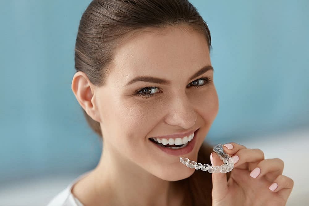 A person smiling and holding an affordable Invisalign clear aligner near their mouth, set against a serene blue background in San Antonio, TX