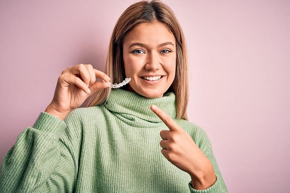 A woman in a green sweater holds and points to a clear dental aligner, smiling against a pink background, showcasing the benefits of an Invisalign Payment Plan in San Antonio, TX