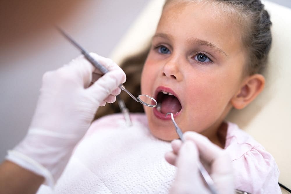 A young girl in a dental chair has her teeth examined by the dentist, who uses a mirror and tool to check for any signs of tongue thrust in San Antonio, TX