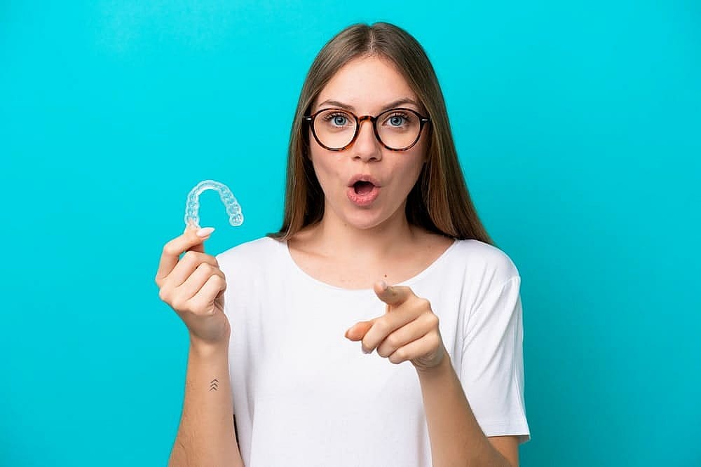 A woman with glasses holds a clear dental retainer, pointing forward with confidence, as she stands against a blue backdrop. Curious if treatments like Invisalign might be covered by insurance in San Antonio, TX