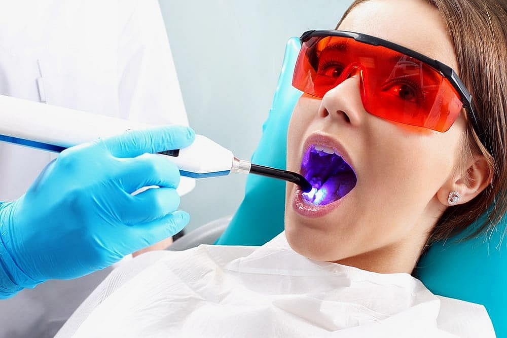 A person undergoing a dental procedure with a curing light, reminiscent of those used in Invisalign vs Braces treatments. They are wearing red protective glasses and a white bib, while the dentist carefully holds the light device in San Antonio, TX