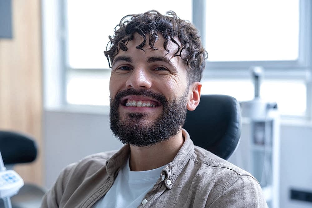 A smiling man with clear braces near me and a beard sits comfortably in a dental chair, sunlight streaming through the window behind him in San Antonio, TX