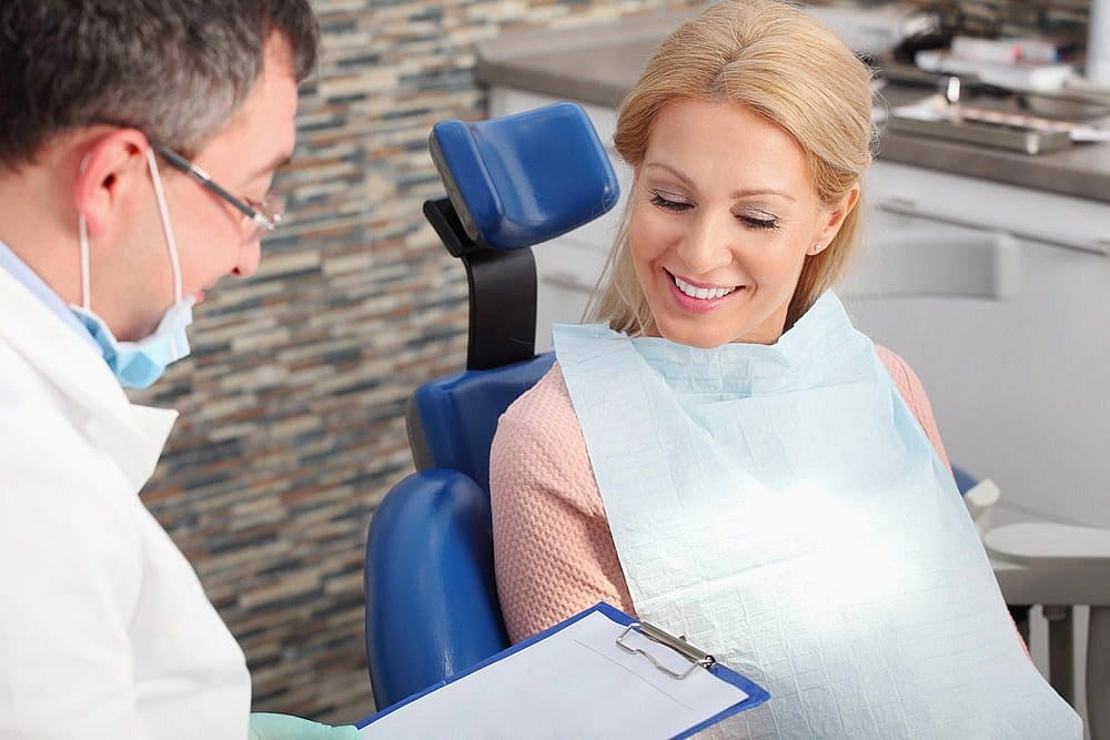 A dentist discusses Invisalign insurance coverage with a smiling patient in the modern room, featuring sleek stone wall panels. The patient sits comfortably in the dental chair, intrigued by the clipboard details in San Antonio, TX