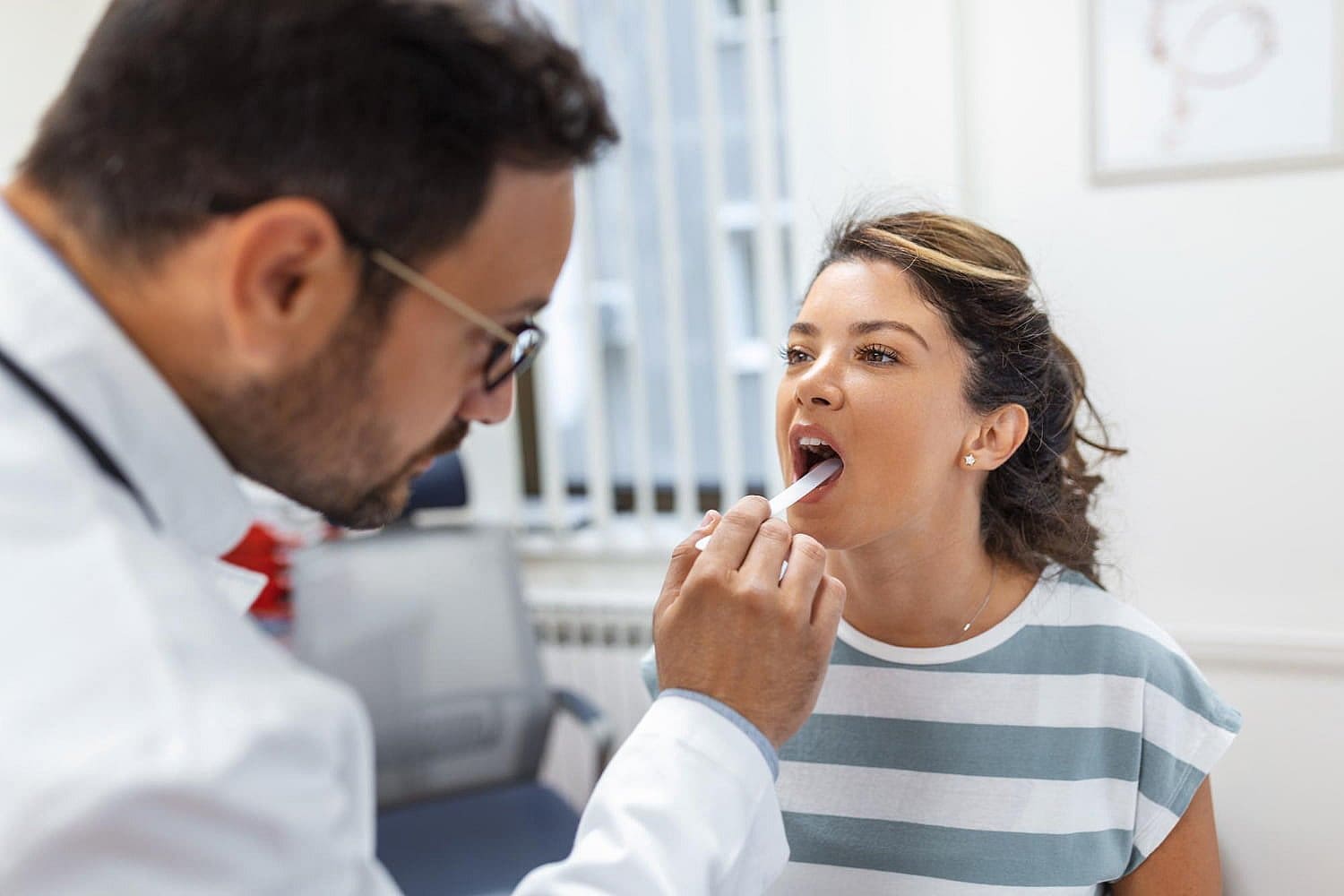 In a medical office, a doctor examines a woman's throat using a tongue depressor, paying special attention to potential signs of tongue thrust in San Antonio, TX