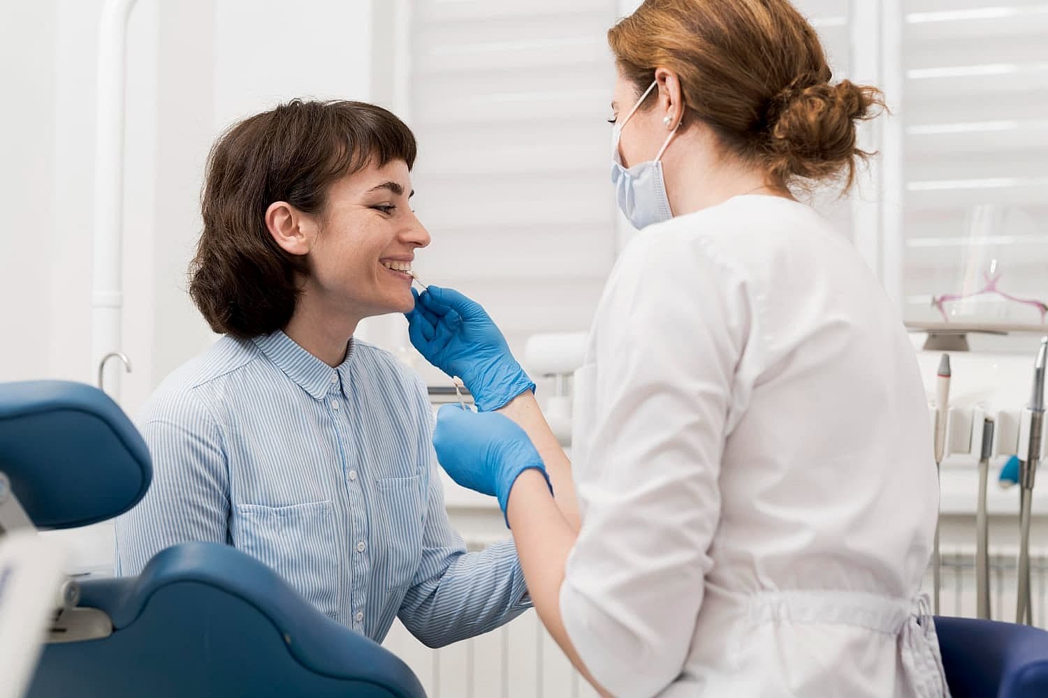 A dentist wearing gloves examines a smiling patient's teeth in a dental clinic, discussing the benefits and flexibility of an Invisalign payment plan in San Antonio, TX