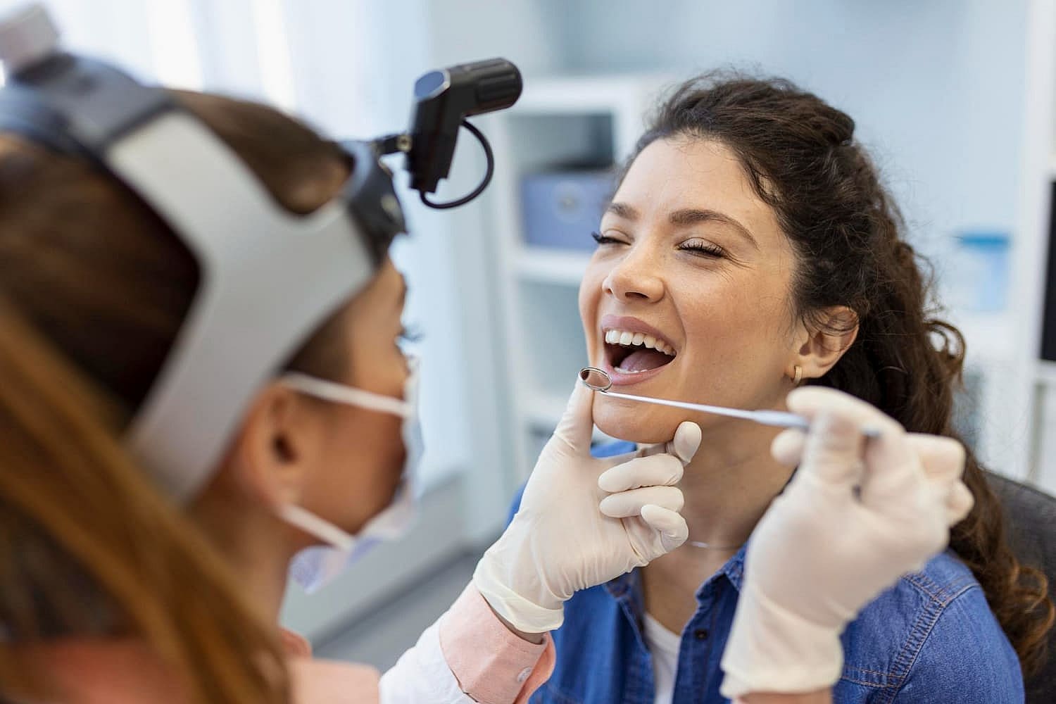 A healthcare professional examines a woman's throat using a tongue depressor and light in a medical office. Both are wearing masks, as the examiner, equipped with a head-mounted light, checks for signs typically seen in mouth breathers in San Antonio, TX