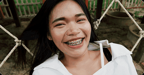 A person with long hair and braces, showcasing the benefits of adult orthodontics, smiles while sitting on a hammock in a white shirt in San Antonio, TX