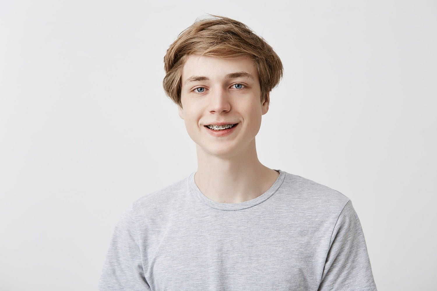 A young person with a cheerful smile and braces beams near me confidently, their short hair complementing the casual gray T-shirt against the plain white background in San Antonio, TX