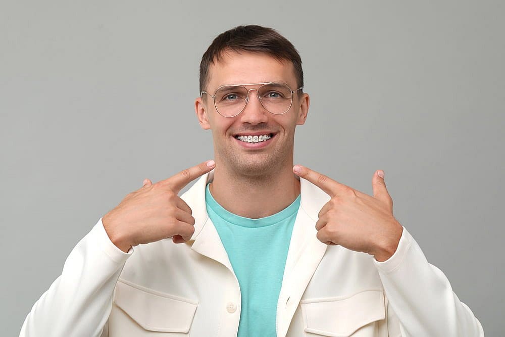 A man with glasses and braces smiles, pointing at his adult orthodontics. He wears a light blue shirt and a cream jacket, all set against a gray background in San Antonio, TX