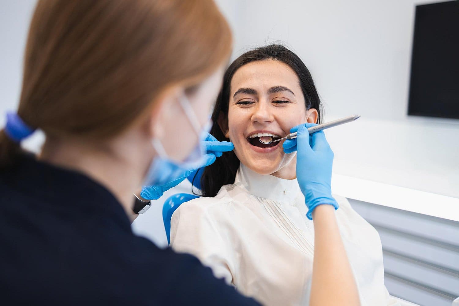 A dentist wearing blue gloves examines a smiling woman's teeth with a dental mirror and probe, carefully checking for signs of tongue thrust. The woman is seated in the dental chair, both focused on the procedure in San Antonio, TX