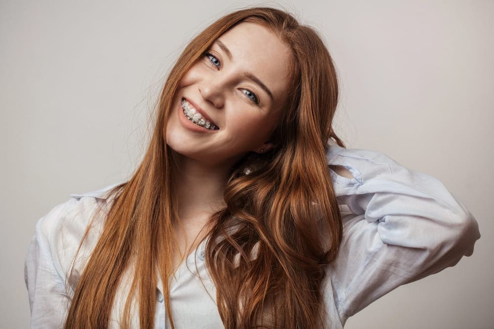 A person with long red hair and clear braces smiles warmly, wearing a light blue shirt, with one hand playfully tucked behind their head in San Antonio, TX