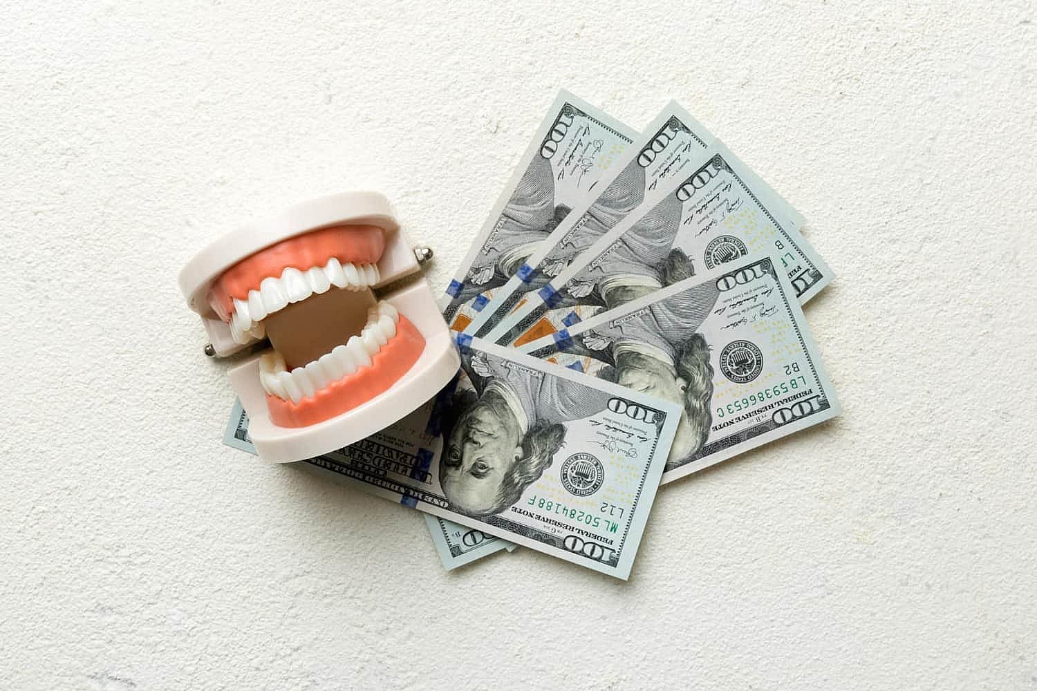 A set of artificial teeth, wearing clear braces, positioned open on top of several scattered $100 bills, all placed on a textured white surface in San Antonio, TX