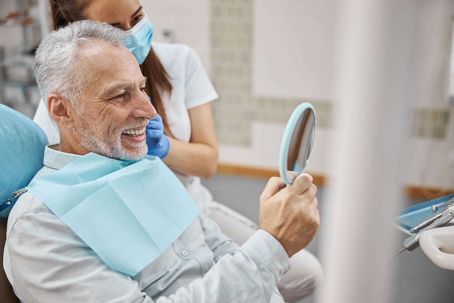 An older man grins, clutching a mirror while seated in a dental chair, reflecting on his journey with adult orthodontics. Beside him, a masked and gloved female dental professional stands proudly in San Antonio, TX
