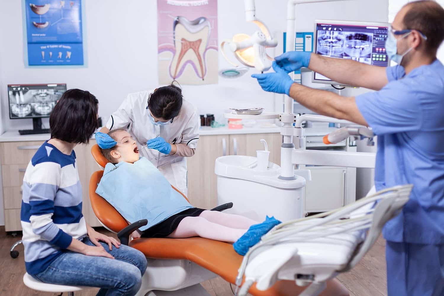 A child with metal braces sits in a dental chair, being examined by the dentist, while a parent watches nearby. Another dental professional adjusts the overhead light for better visibility in San Antonio, TX