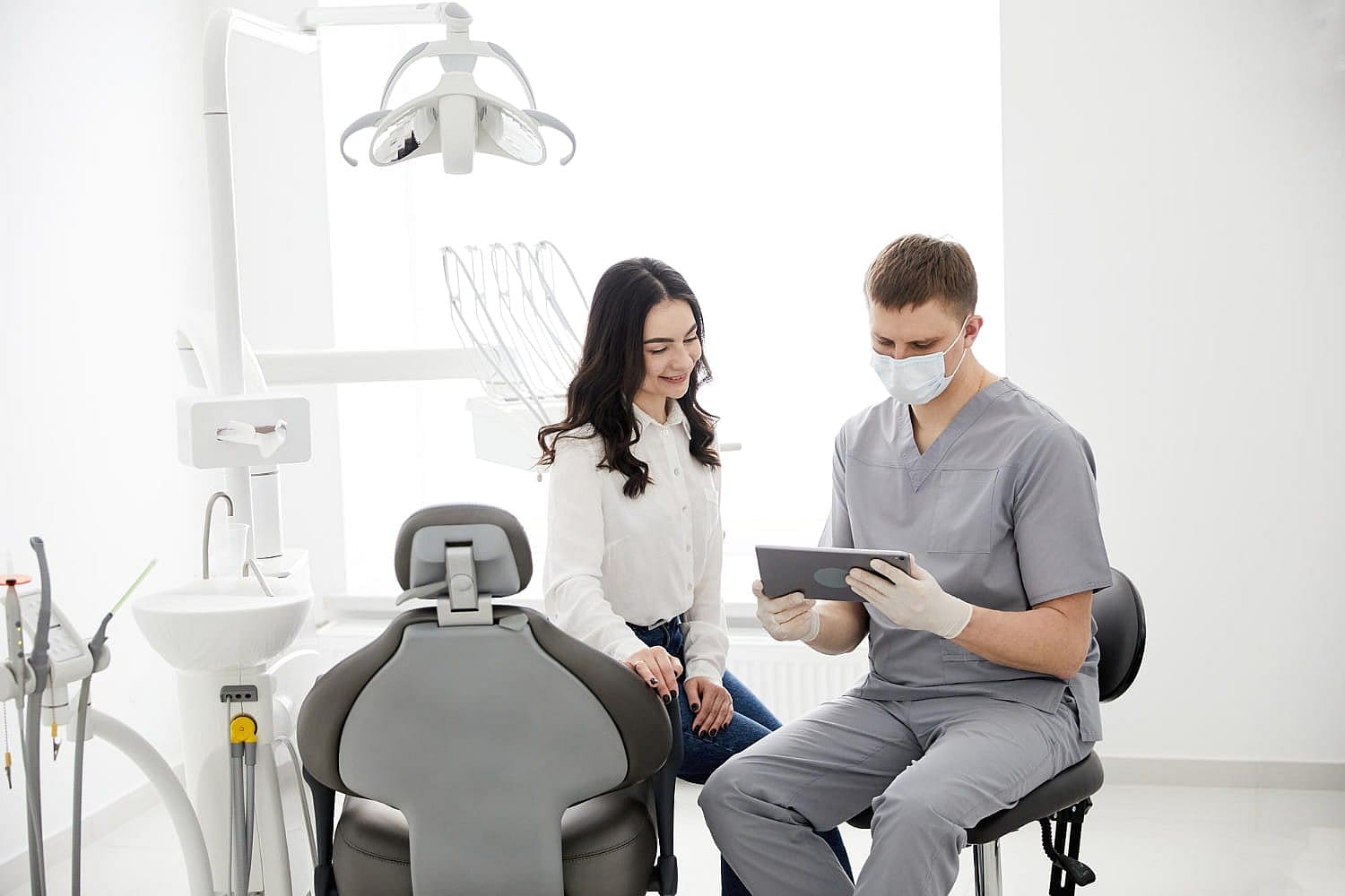 A dentist in scrubs and mask shows a tablet to a seated woman in an examination room, discussing tongue thrust issues, with dental equipment subtly arranged in the background in San Antonio, TX