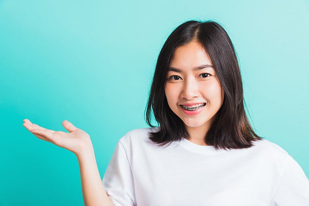 A woman with medium-length hair and metal braces smiles, standing against a turquoise background, wearing a white t-shirt. Her left hand is raised in a presenting gesture in San Antonio, TX