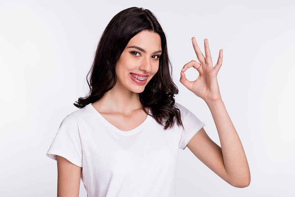 A woman with long dark hair, showcasing the success of adult orthodontics, smiles brightly in a white t-shirt and braces, making an "OK" gesture against a plain background in San Antonio, TX