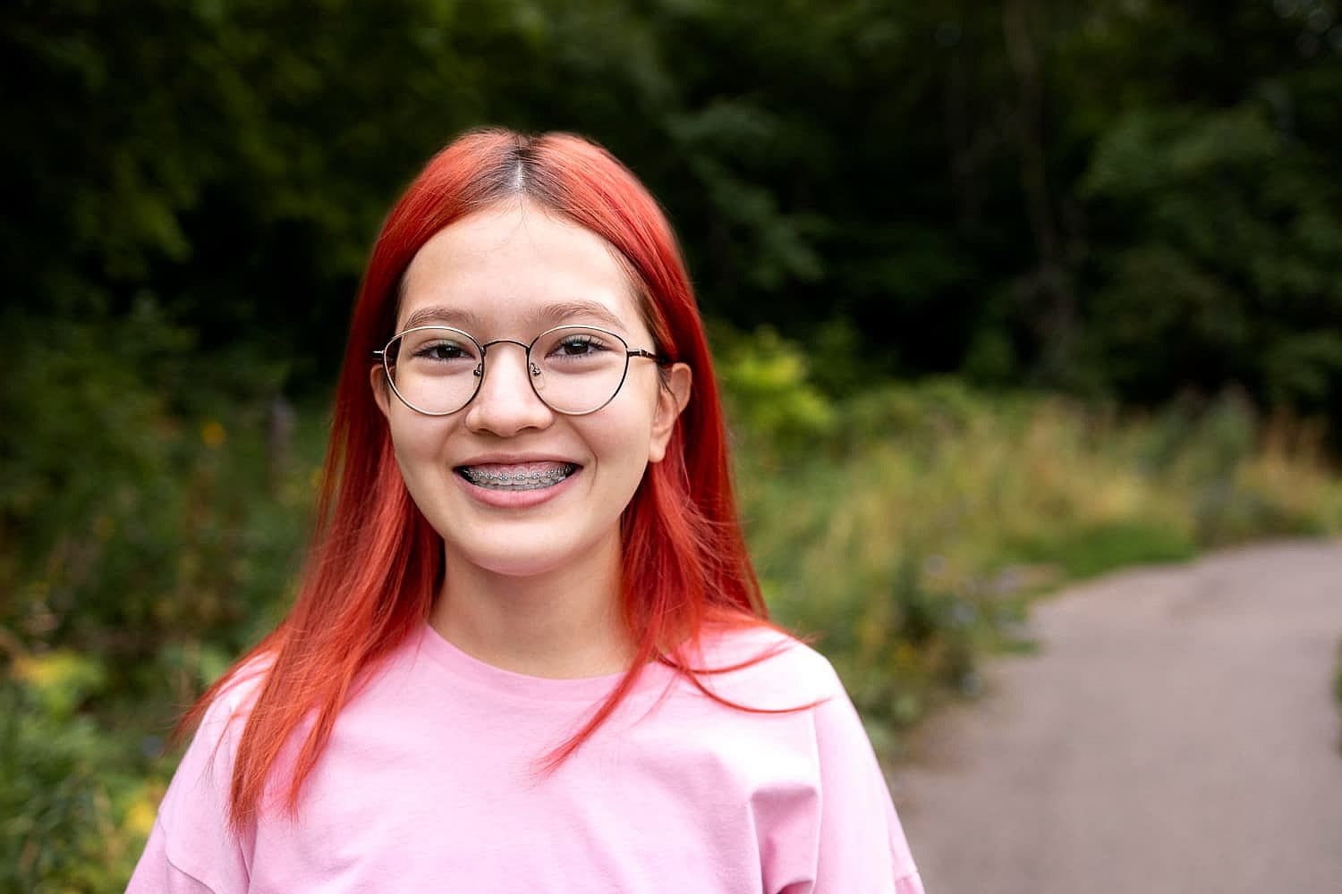 A person with red hair, wearing glasses and braces that subtly correct an underbite, smiles while standing on a path in a green park, dressed in a cheerful pink shirt in San Antonio, TX