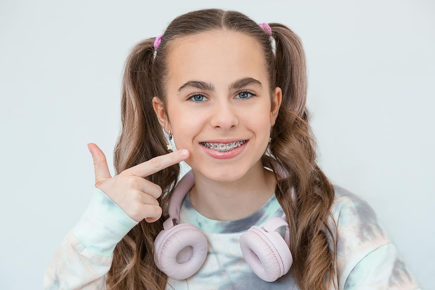 A teen girl with long hair in pigtails, wearing a tie-dye shirt, points at her mouth with one hand while pink headphones rest around her neck. Her smile highlights the classic debate: Invisalign vs. braces in San Antonio, TX