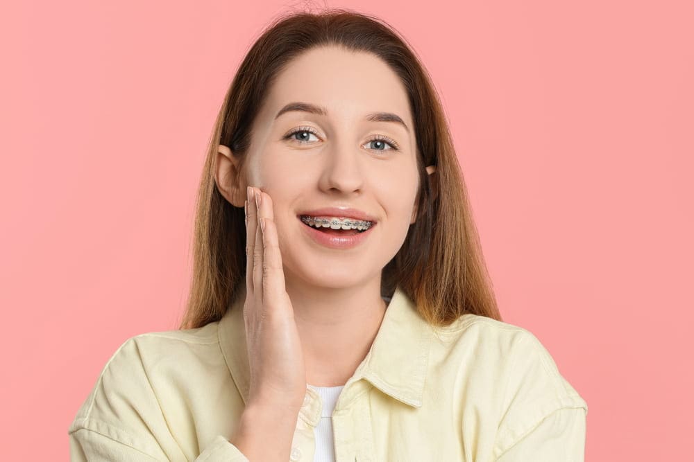 A young woman with long brown hair and metal braces, courtesy of her San Antonio, TX orthodontist, smiles while touching her cheek. She is wearing a light yellow shirt against a pink background, confidently showcasing her metal braces.