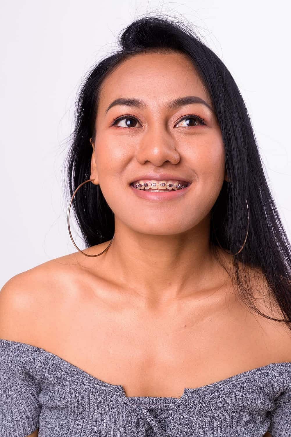 Woman with long black hair, wearing hoop earrings and a gray off-shoulder top, smiling confidently. Her adult braces add charm as she gazes upward in San Antonio, TX