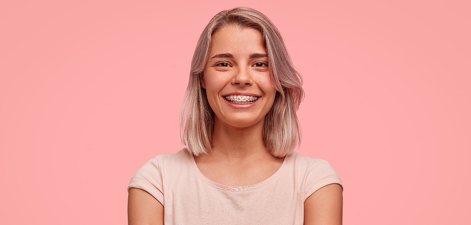 A woman with braces near me beams brightly, donning a light pink shirt against a vibrant pink background in San Antonio, TX