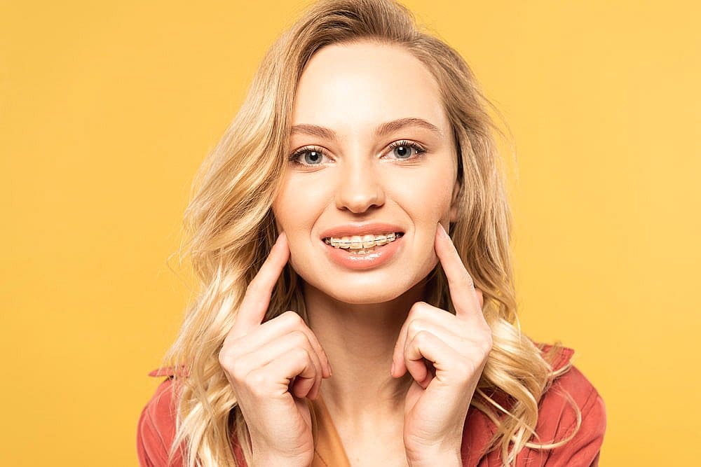Fiesta Orthodontics A woman with long blonde hair smiles, showcasing her clear braces while pointing at them with both index fingers. She stands against a solid yellow background, confidently wearing a red jacket in San Antonio, TX