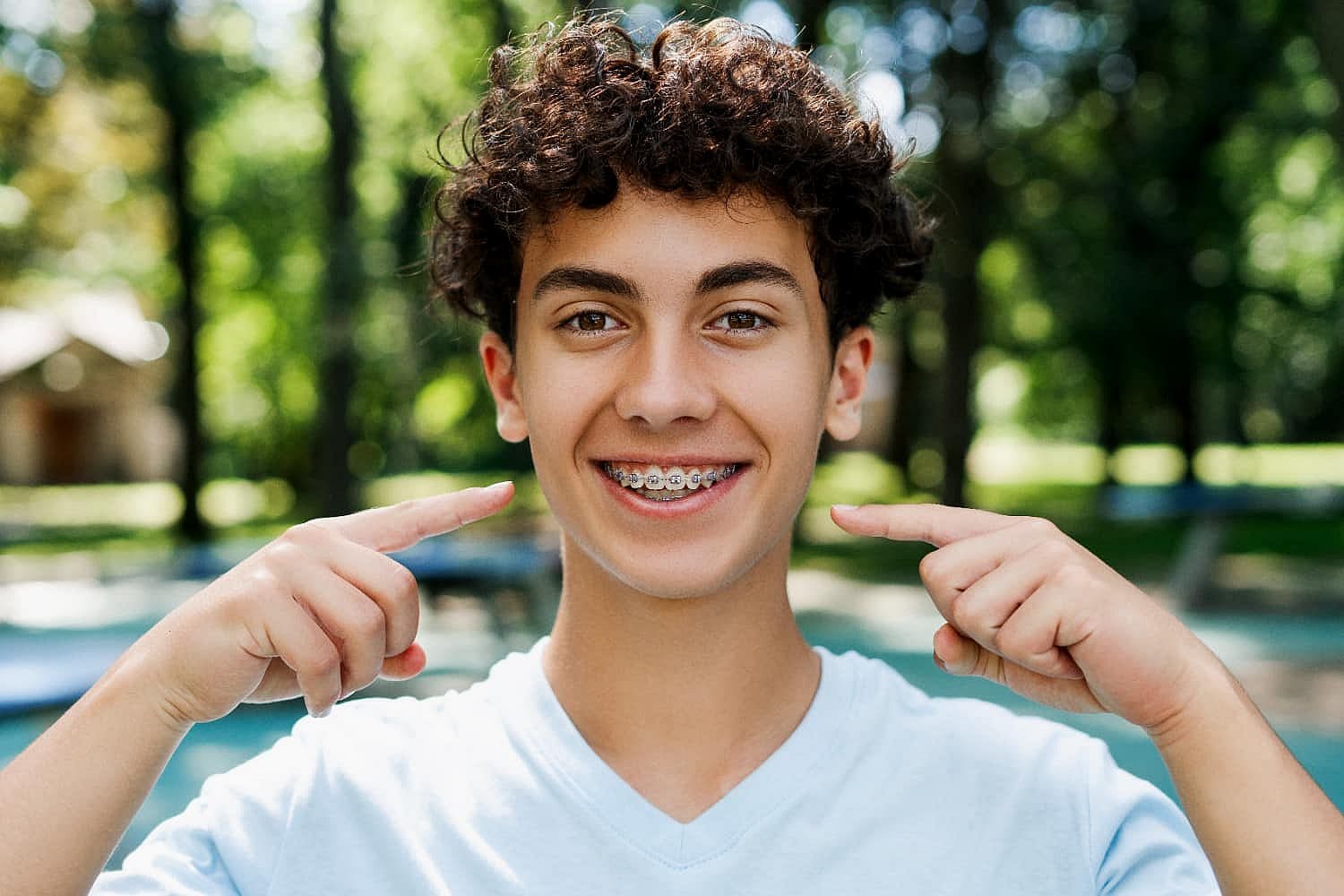 Teen boy outdoors, flashing a bright smile with braces, proudly pointing at his teeth, confident they were worth the cost in San Antonio, TX