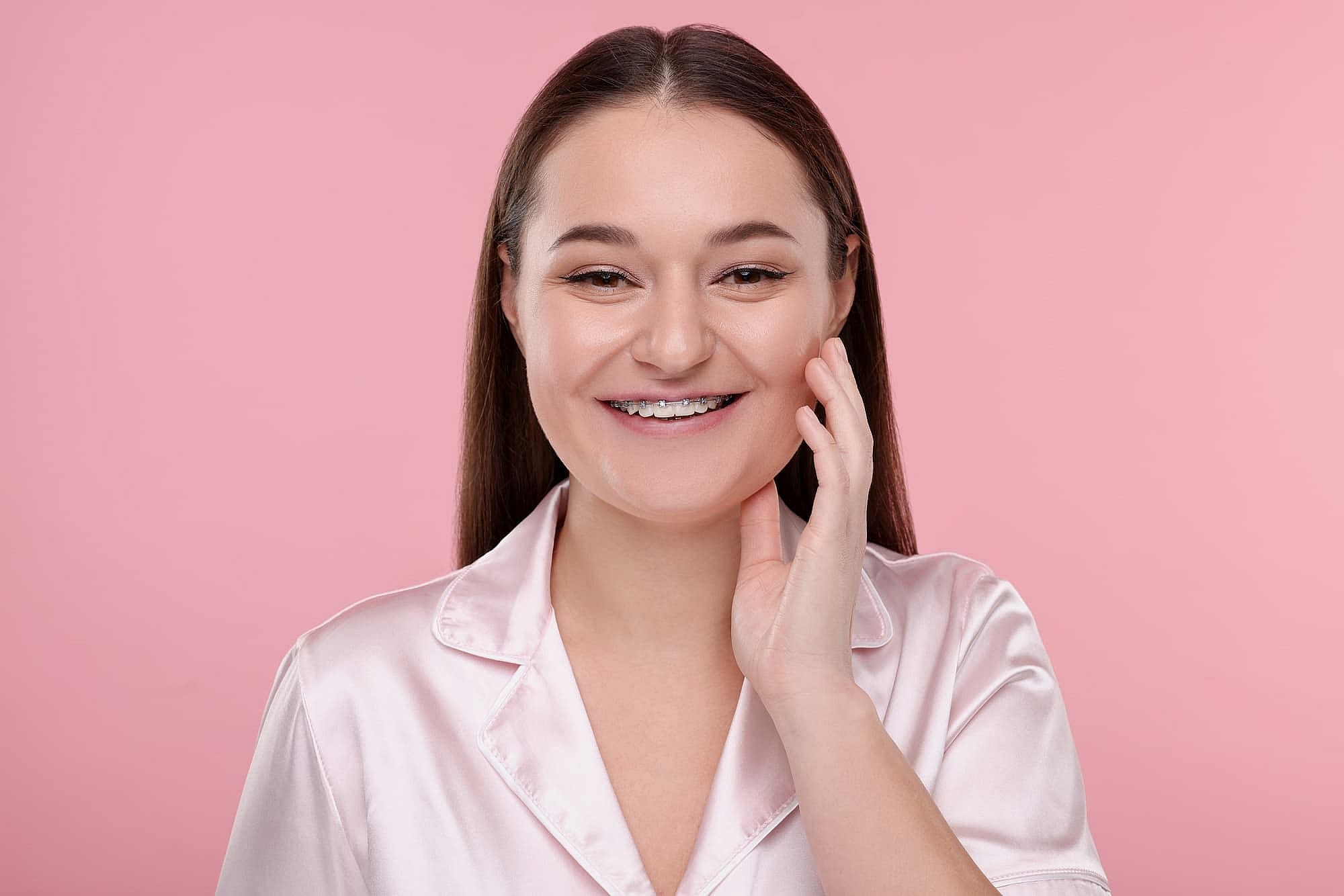 A smiling person with long hair and metal braces touches their cheek. They wear a satin shirt, standing against a pink background in San Antonio, TX