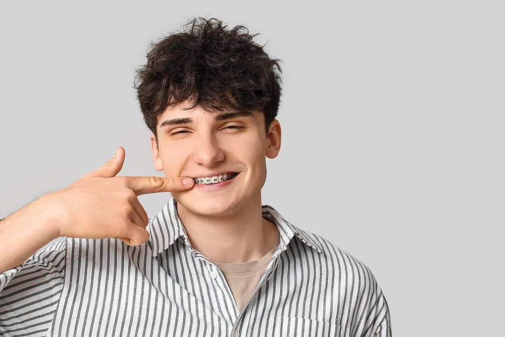 A young man with curly hair points to his adult braces while smiling. He is wearing a striped shirt against a plain background in San Antonio, TX